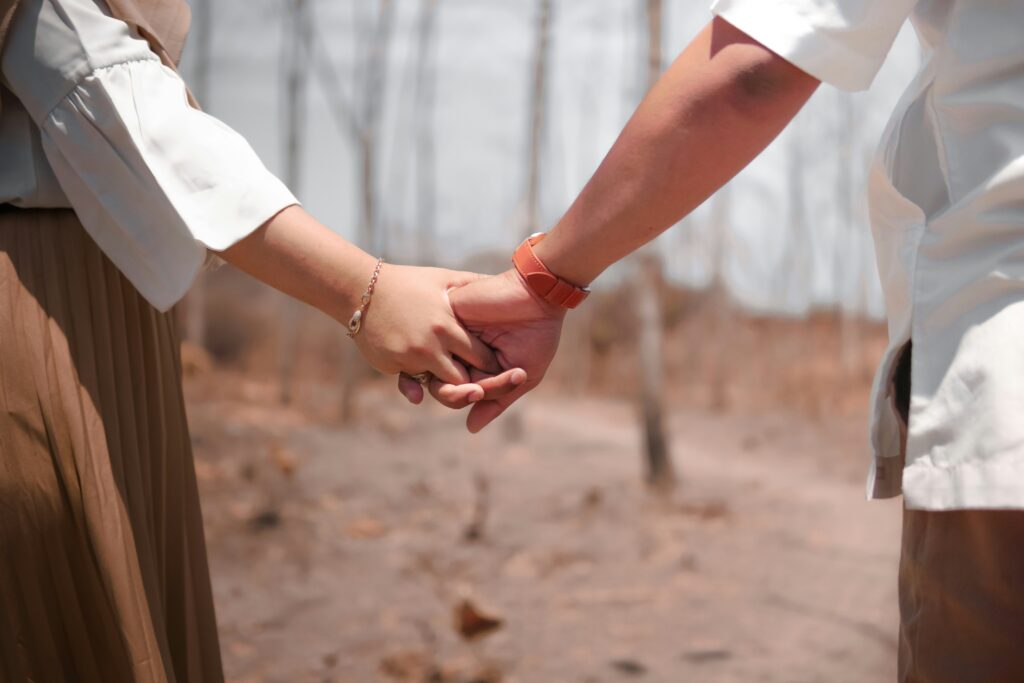 Two people hold hands outdoors in a sunny, dry setting; one wears a brown skirt and bracelet, the other a light outfit.