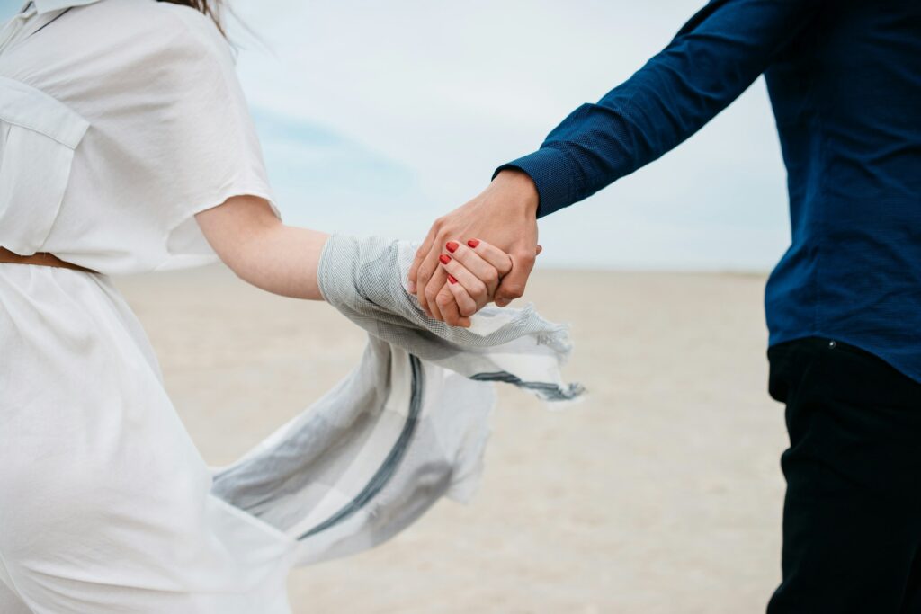 Two people hold hands at the beach, one in a white dress and the other in a dark blue shirt, with a sandy shore in view.